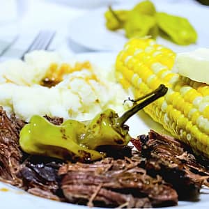 Easy Crockpot Dinner, Mississippi Roast. On a white plate with a pepperoncini pepper, corn on the cob with a butter slice on top, and mashed potatoes with Mississippi gravy