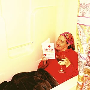 Photo of Michelle Grewe in bath tub wearing a red sweater, a rose hair band, and covered in a brown blanket, reading book Mom for the Holidays while sipping red wine in a wine glass