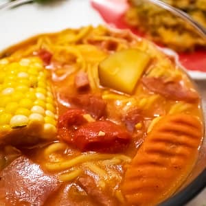 Puerto Rican Ham and Salami Soup featuring corn on the cob, salami, carrots, noodles, and red peppers