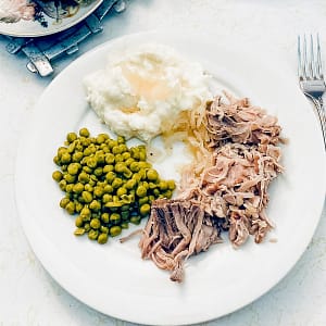 Photo of Pork Roast with Sauerkraut, mashed potatoes and peas on a white plate on a white tabletop.