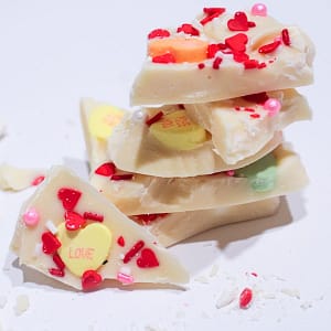 White Chocolate Sweethearts Bark. 5 Chunks of bark stacked with one chunk facing front leaning on stack showing yellow conversation heart that says, love, and red heart sprinkles. with some shaved chocolate and sprinkles around it on a white table with a gray background.