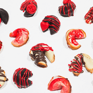 Photo of fortune cookies covered in drippy gooey milk chocolate and red chocolate foe shizzle drizzles on a cooling rack on a white background.